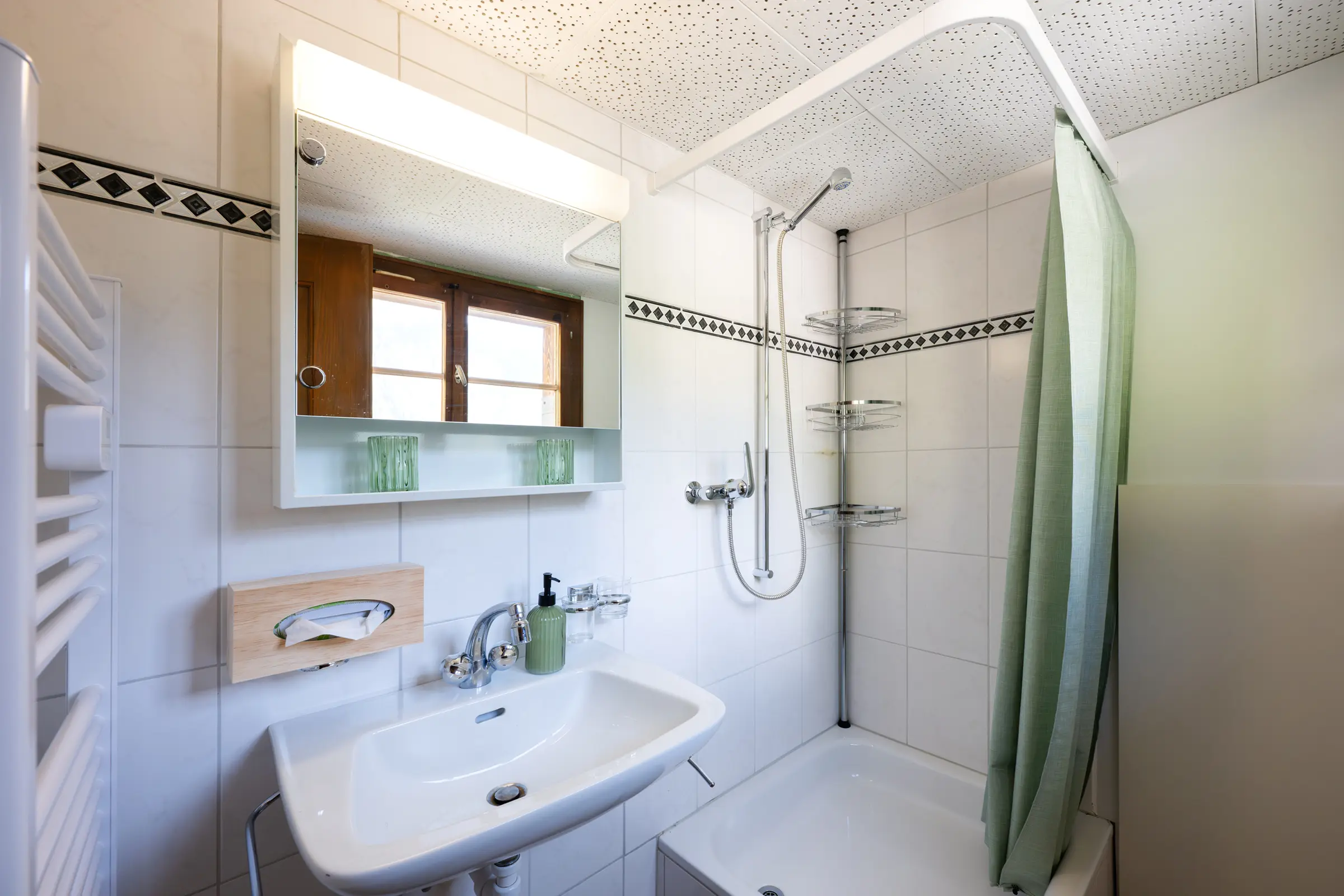 Bathroom with a basin, mirror cabinet and towel radiator on the left, on the right a shower over the bath with a green shower curtain, white wall tiles with a dark border.