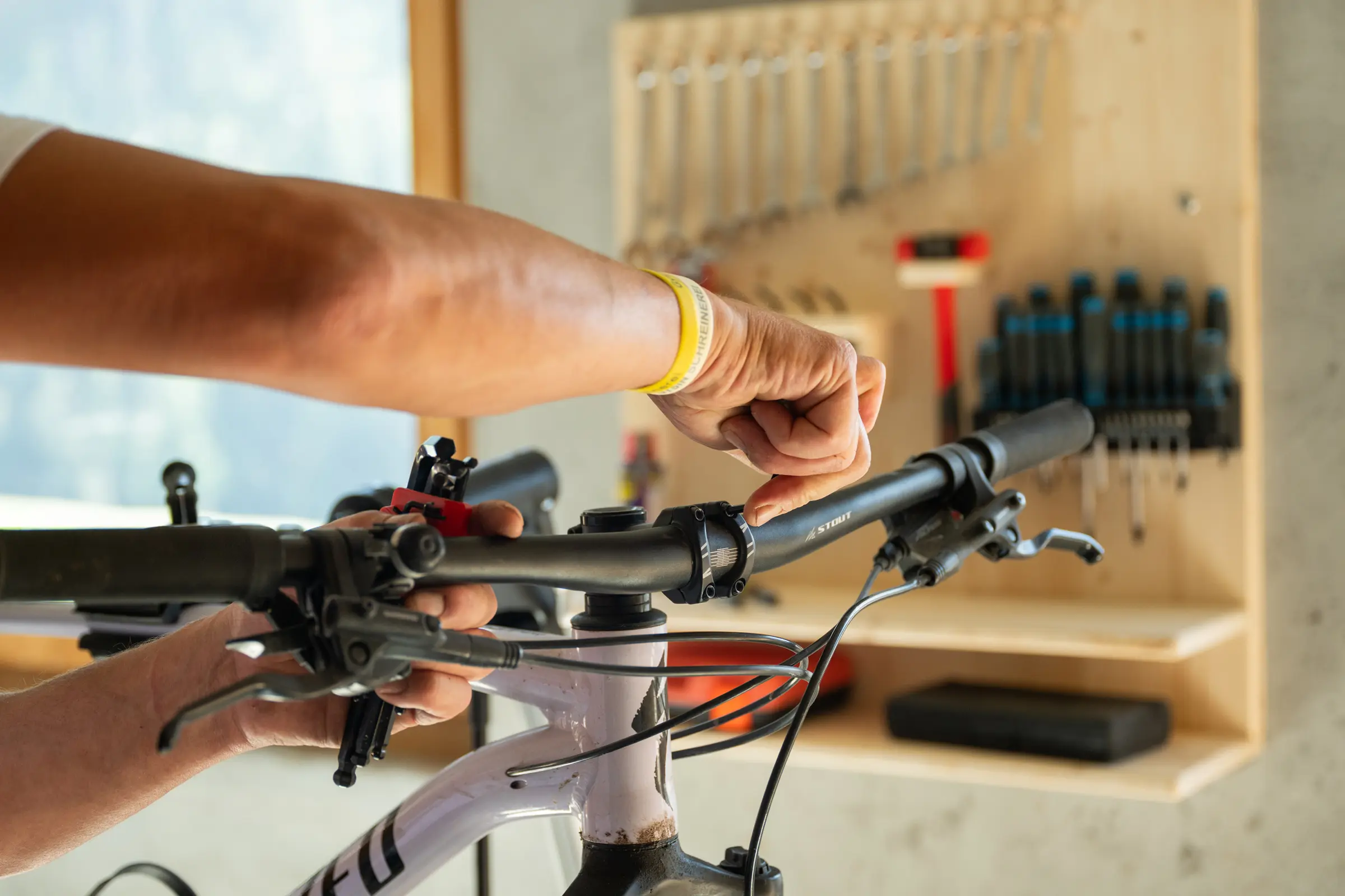 Two hands adjusting the handlebars of a mountain bike, the tool board with spanners and screwdrivers blurred behind them.