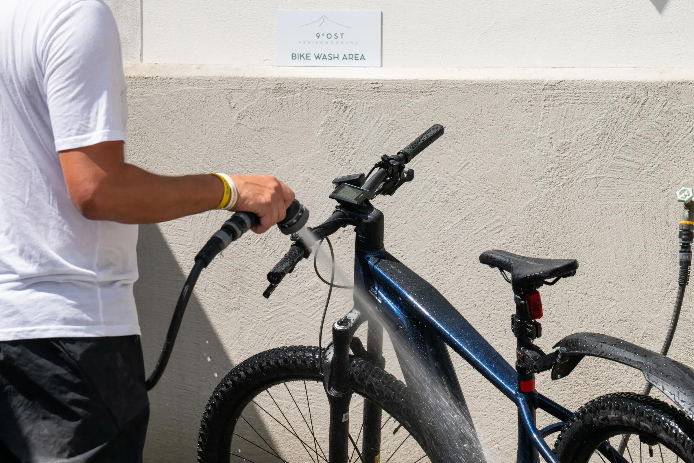 Person hosing down a mountain bike, on the wall the Bike Wash Area sign with the mark of 9 Grad Ost.