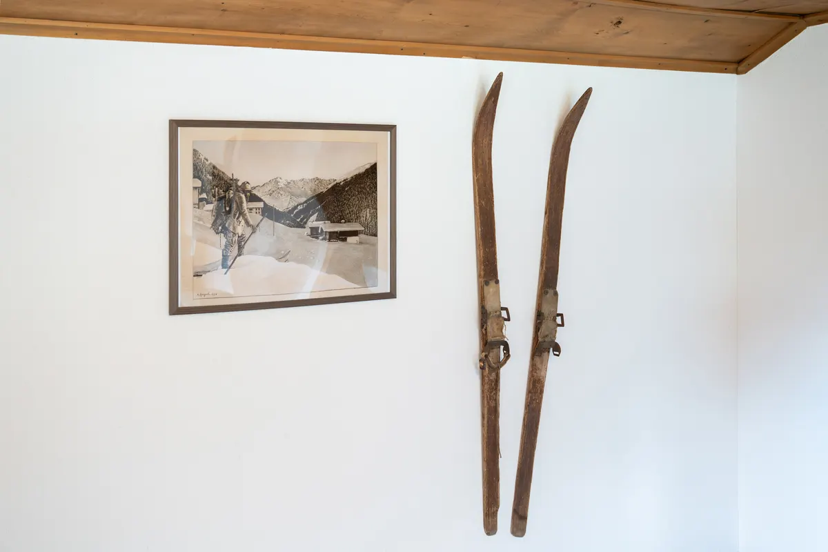 Two old wooden skis with leather bindings on a white wall, on the left a framed old photograph of a skier in front of mountains and houses.
