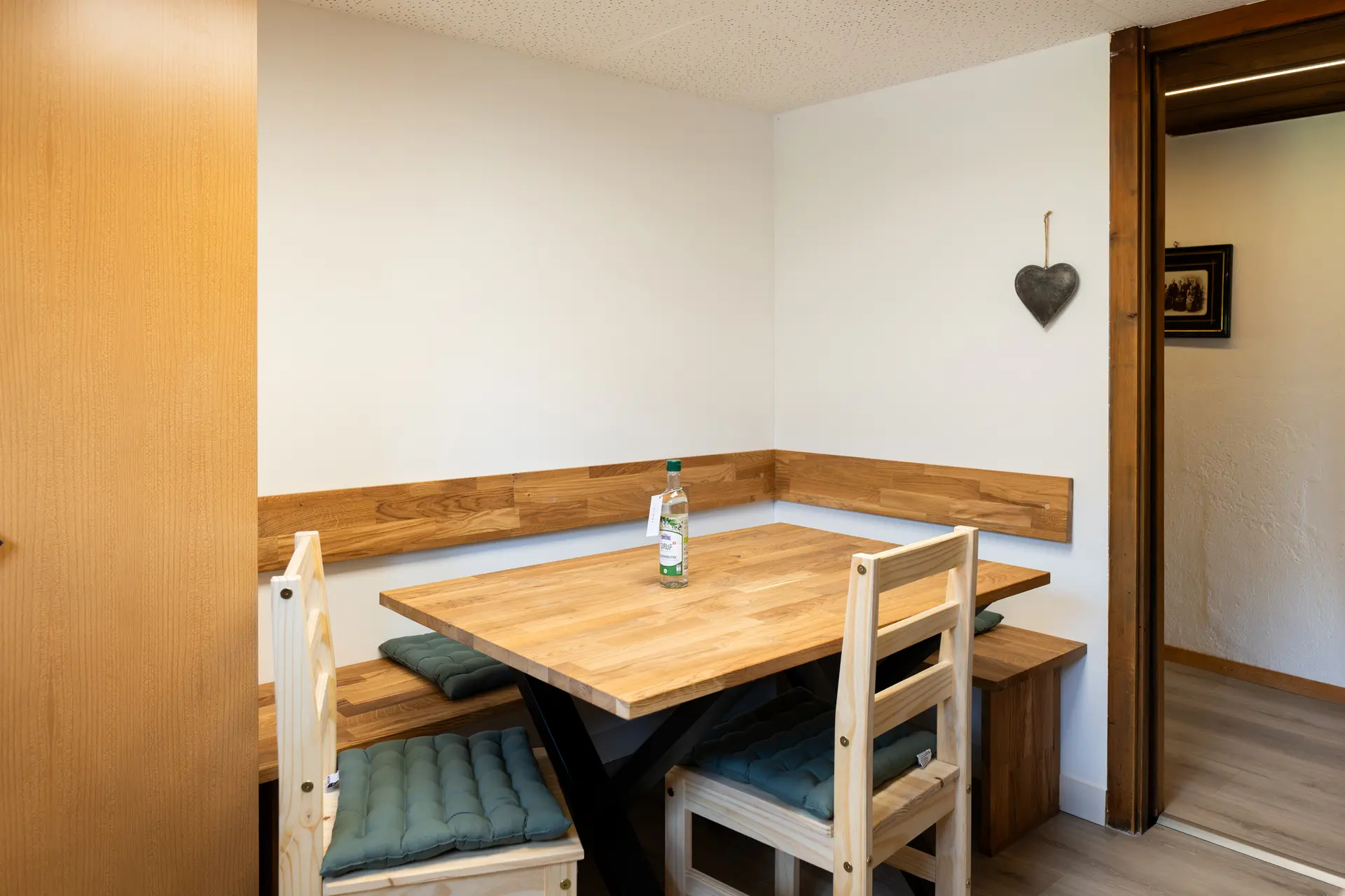 Dining area with a wooden table, a corner bench with green seat cushions and two light wooden chairs, a slate heart on the wall, on the right the doorway to the hall.