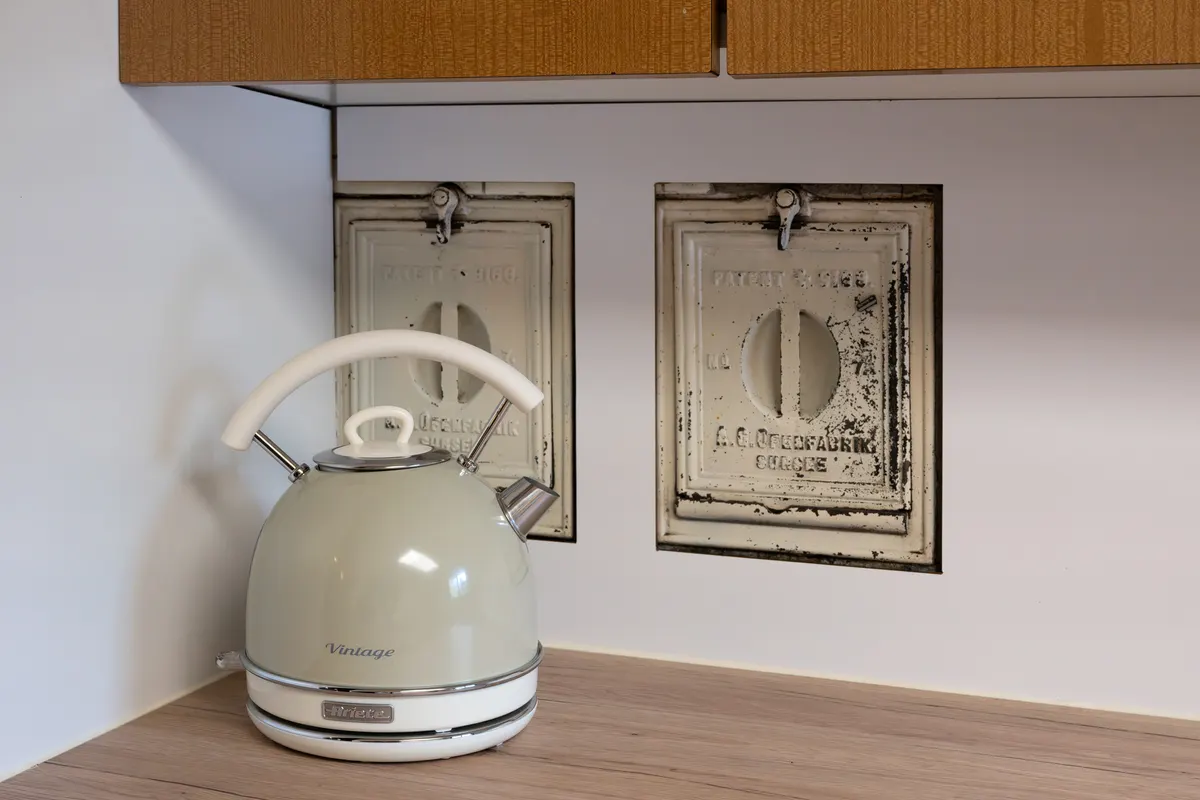 Cream-coloured kettle on the worktop, behind it two old cast-iron stove doors in the wall with embossed lettering.