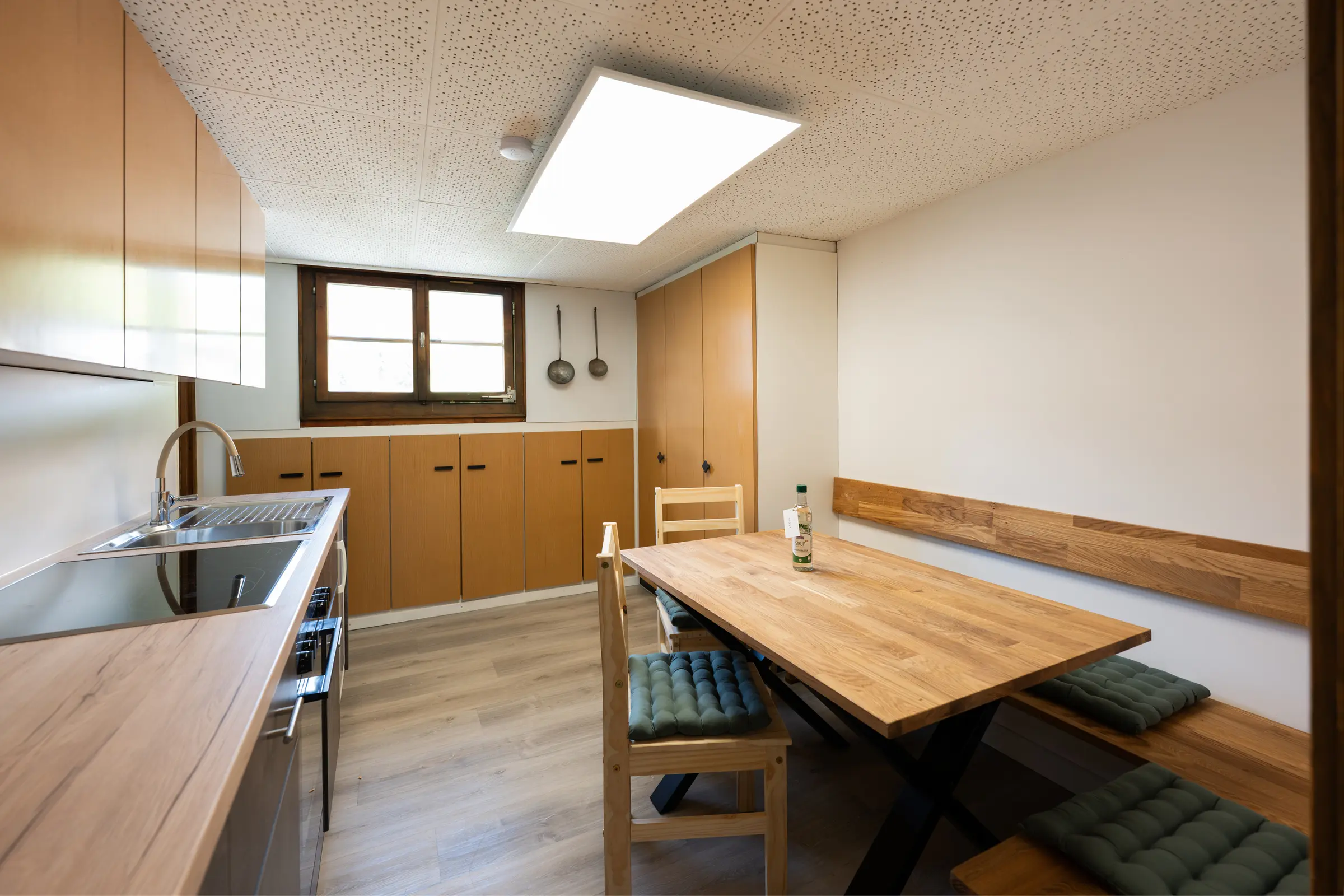 The kitchen from the door: sink and worktop on the left, on the right the wooden dining table with a bench, green seat cushions and a chair, two old ladles hanging on the wall.
