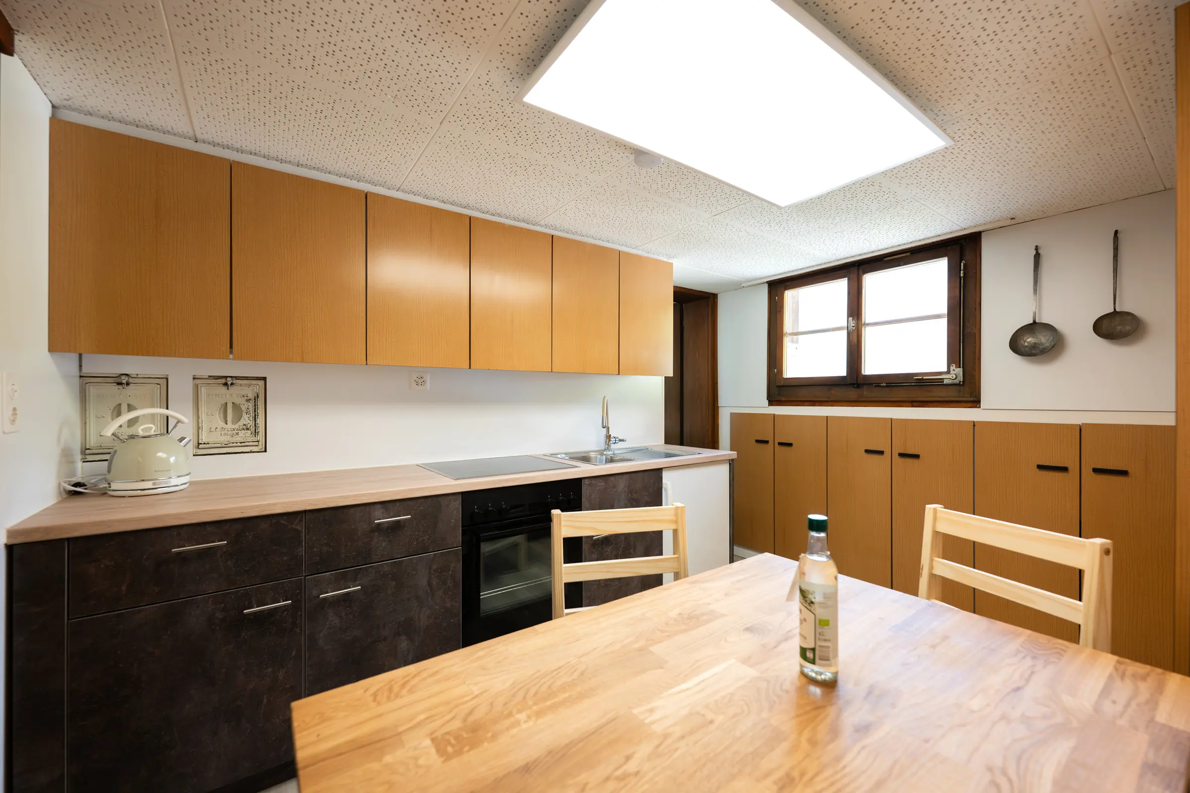 The kitchen units in their full length under the sloping roof, with dark fronts, oven and sink, on the right the wooden dining table, two old ladles on the wall.