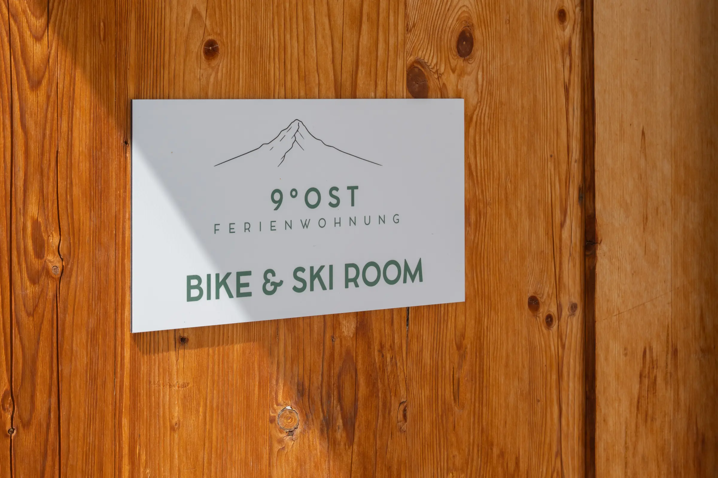 Sign with the mountain mark reading 9 Grad Ost Ferienwohnung, Bike und Ski Room, on a door of Swiss pine.