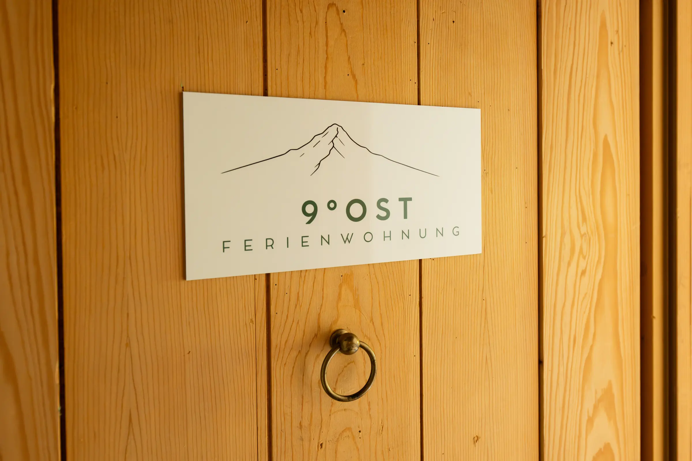 Sign with the mountain mark reading 9 Grad Ost Ferienwohnung on the Swiss pine door of the flat, below it an old brass ring handle.