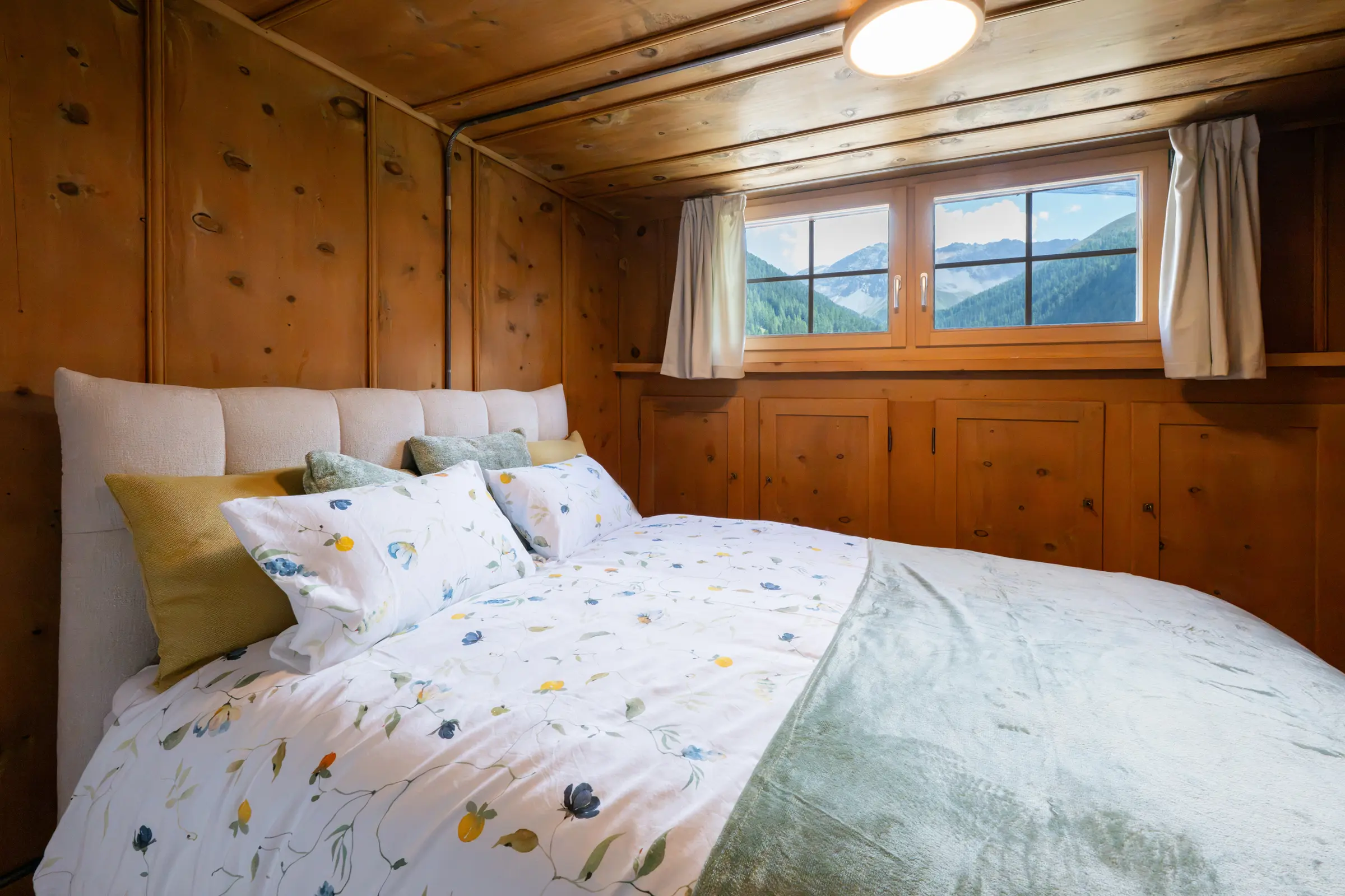 Double bed with a padded headboard and floral bed linen, behind it a window looking onto forest and snowy mountains, old wood panelling with built-in cupboards all around.