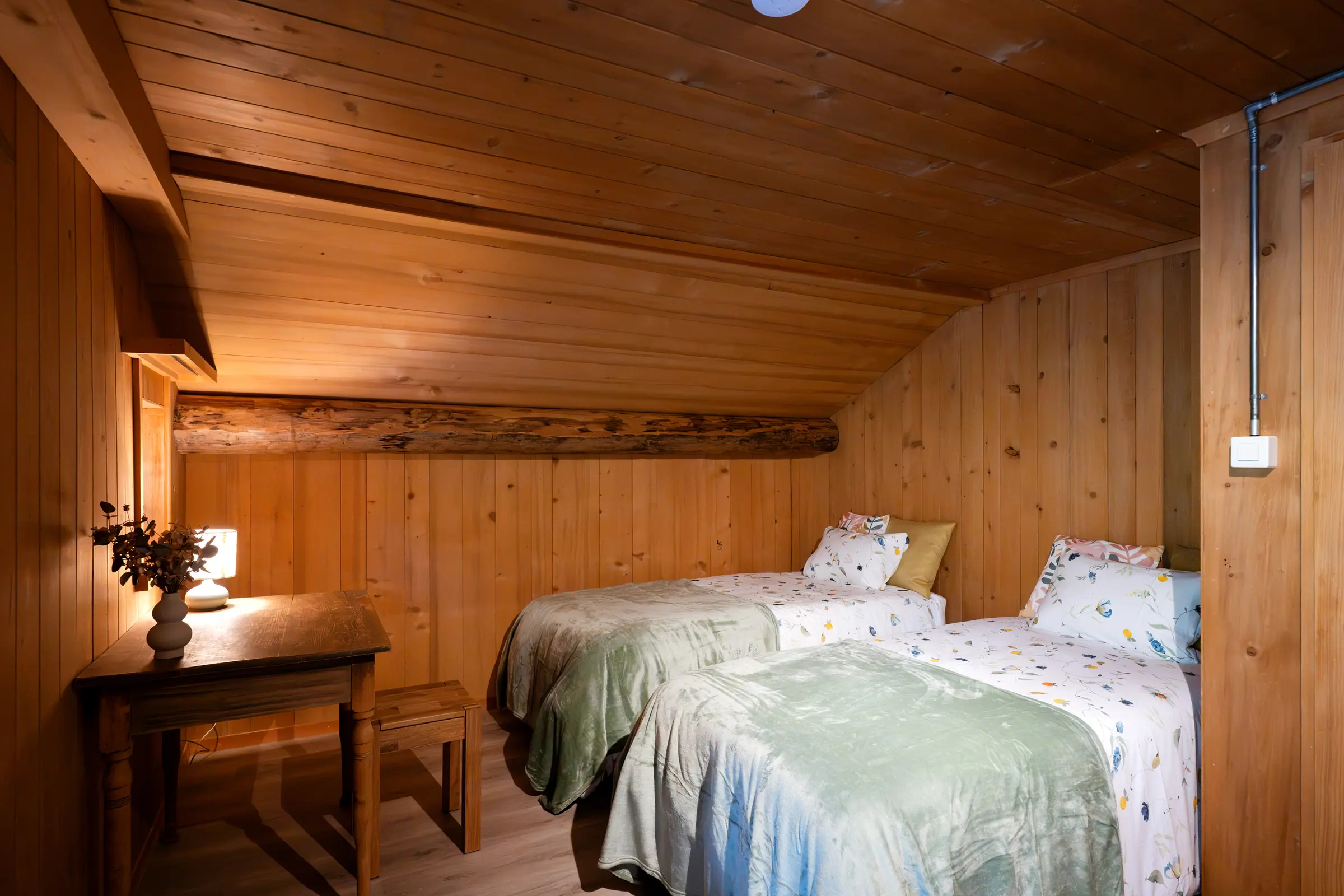 Bedroom under the sloping roof with two single beds, floral bed linen and green bedspreads, on the left an old wooden table with a lamp and dried flowers, an old ceiling beam in the panelling.