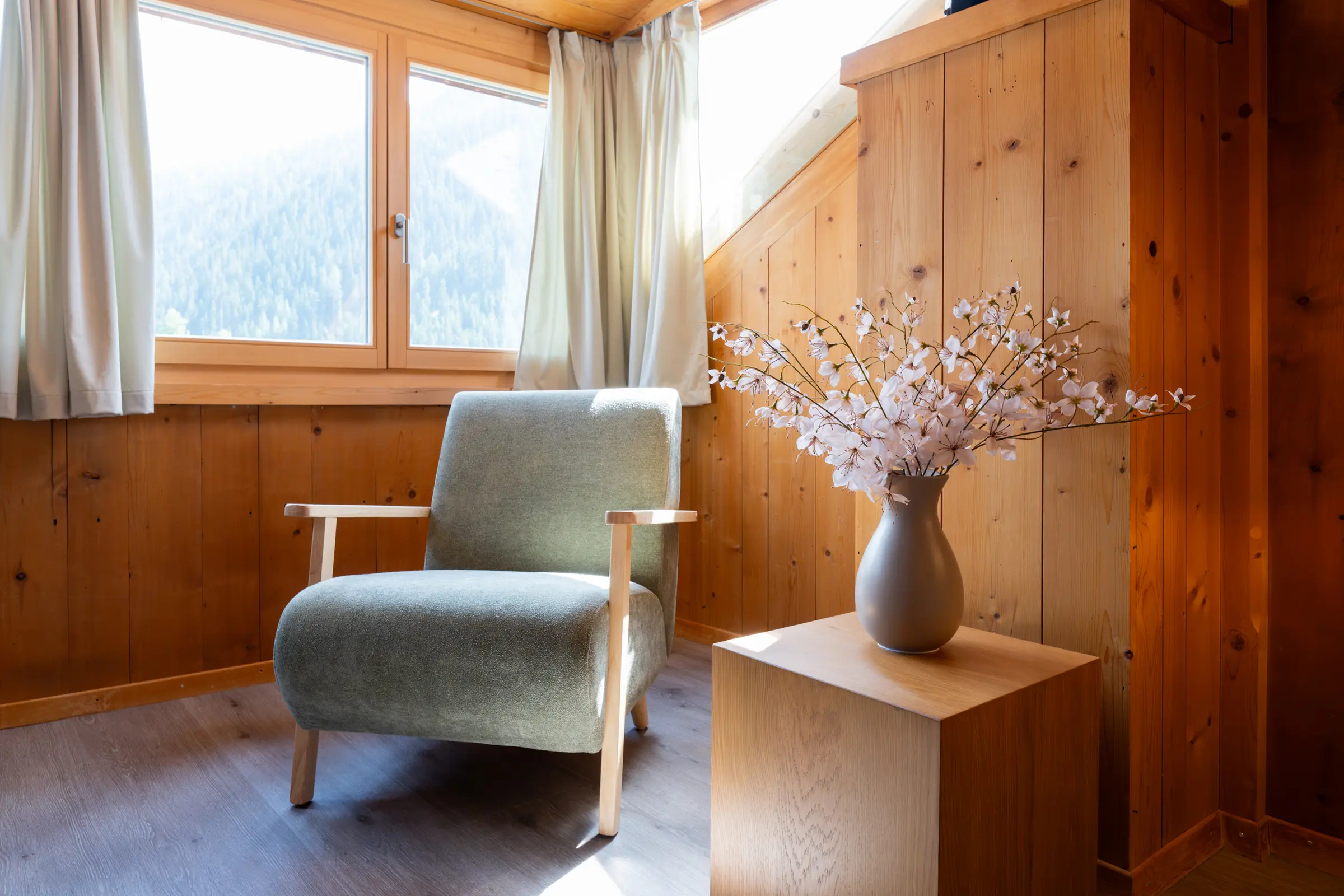 Light green armchair in front of the window with a view of forest and mountains, next to it a vase of flowering branches on a wooden stand, old wood panelling all around.