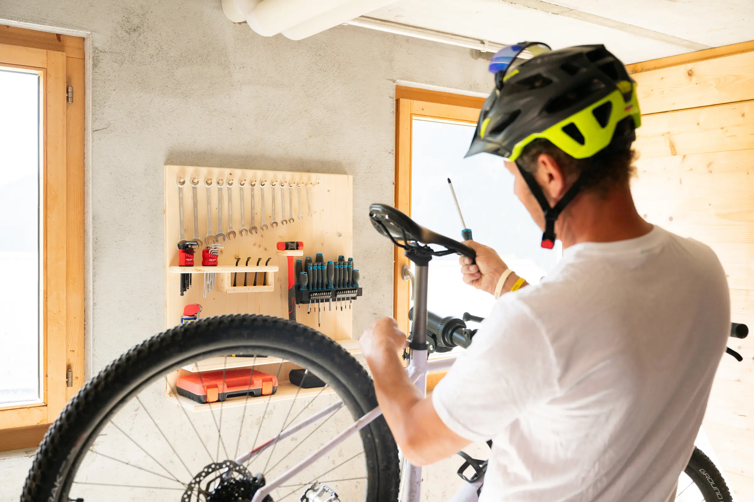 Person in a cycling helmet setting up a mountain bike at the workbench, the tool board with spanners on the wall, a window beside it.