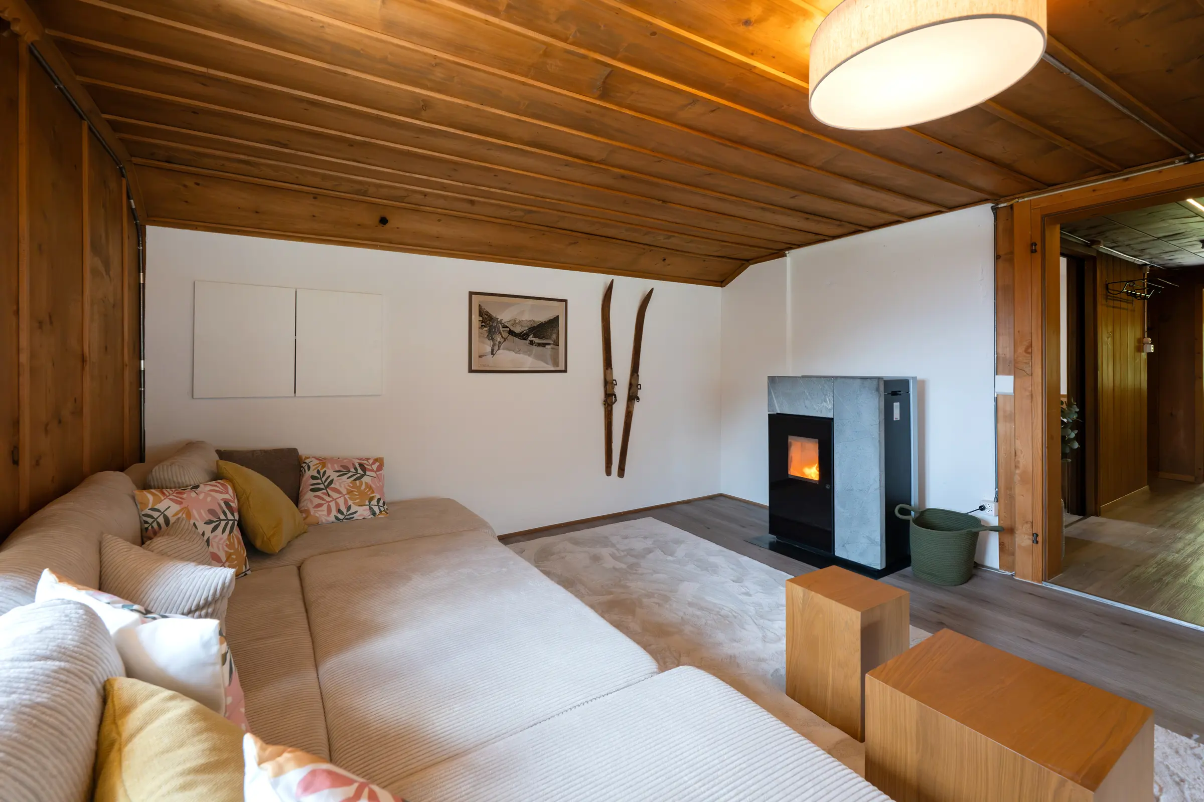 View from the sofa to the pellet stove with a fire burning, beside it two old wooden skis and a framed old photograph on the white wall, on the right the doorway to the hall.